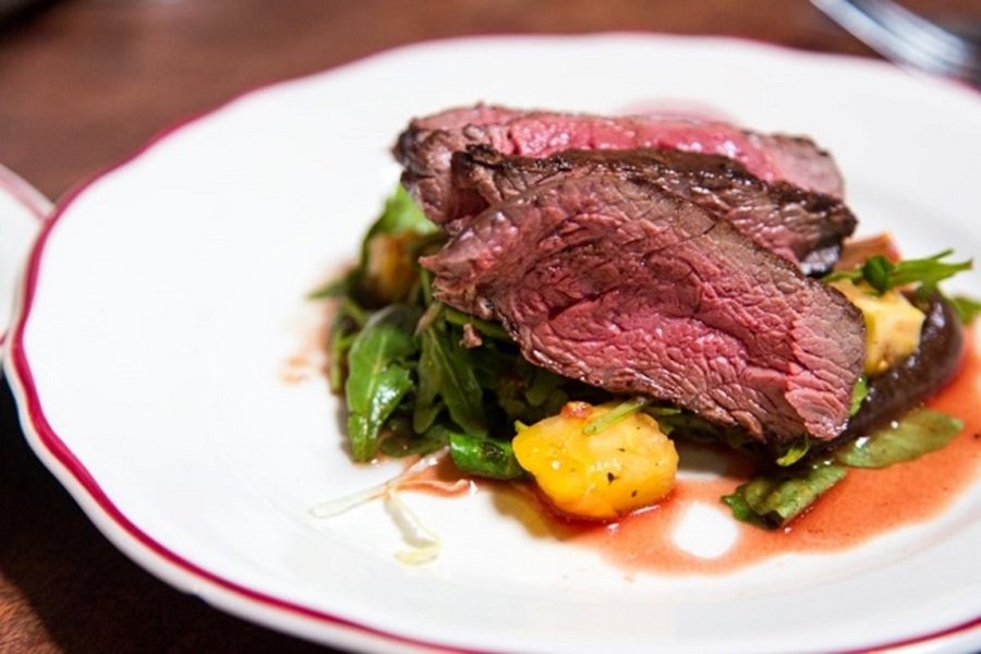 medium-rare steak on a bed of vegetables on a white plate with a wood table in background 