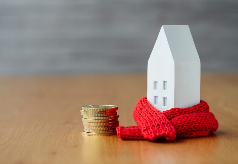 white miniature house wrapped in a red knit scarf next to a stack of coins on a wooden surface with a blurred background