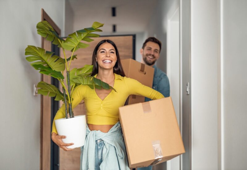 two dark-haired, smiling people carrying boxes and a plant move into a new home confidently after having professionals administer pest control for new homebuyers